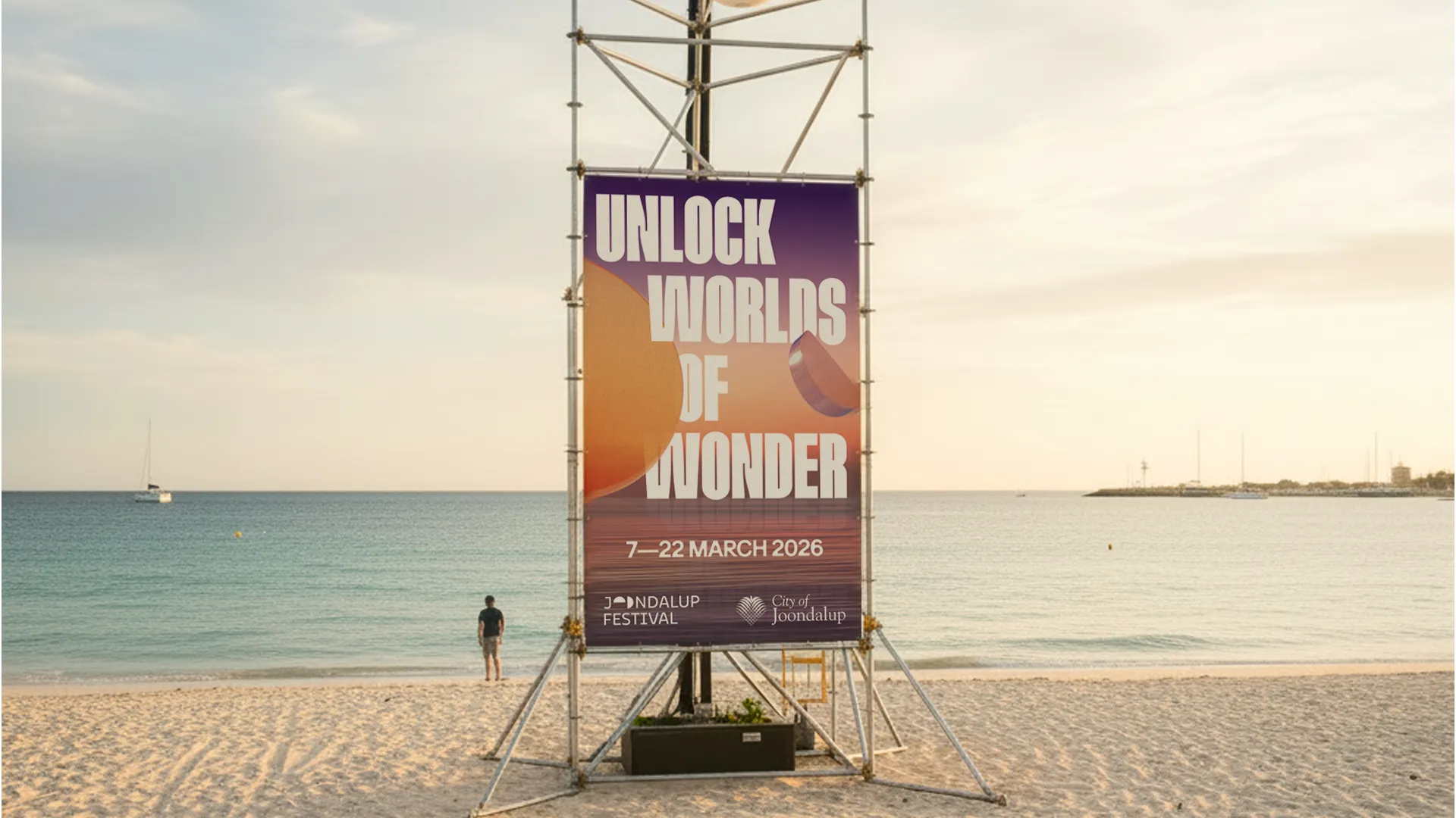Joondalup Festival 2026 poster on beach scaffolding reading Unlock Worlds of Wonder with ocean and boats in the background at golden hour